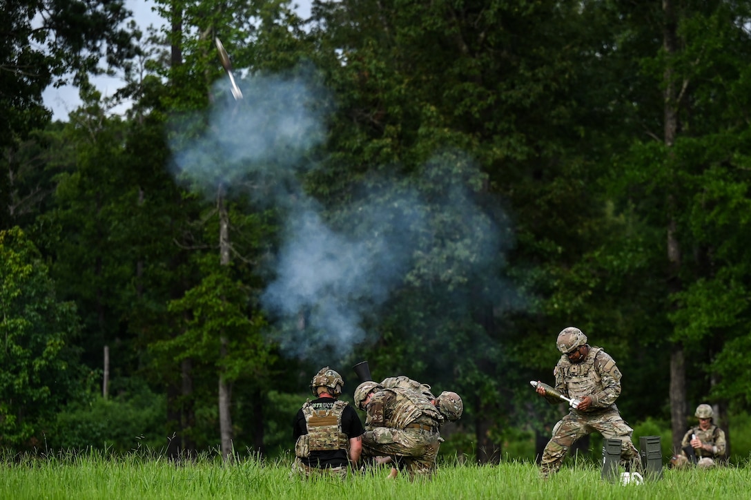 Two soldiers in tactical gear crouch in grass in front of trees to launch a weapon while another holds a small missile.