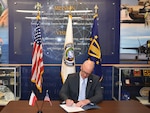 Seated person in a business suit signs a document with flags in the background.
