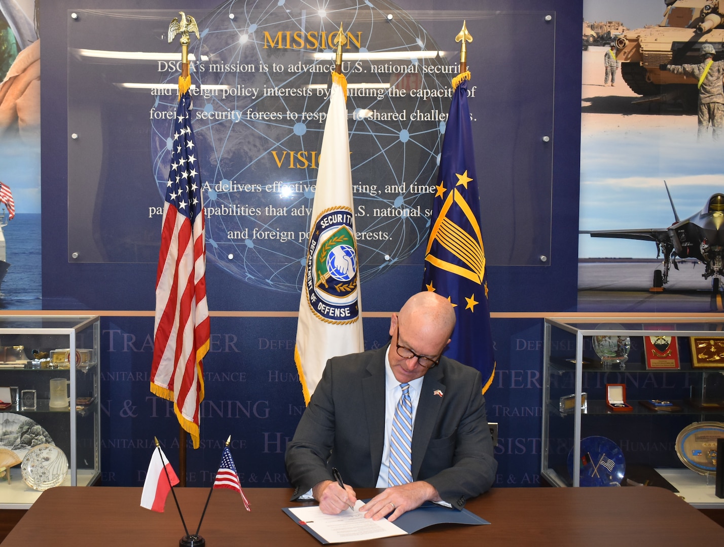 Seated person in a business suit signs a document with flags in the background.