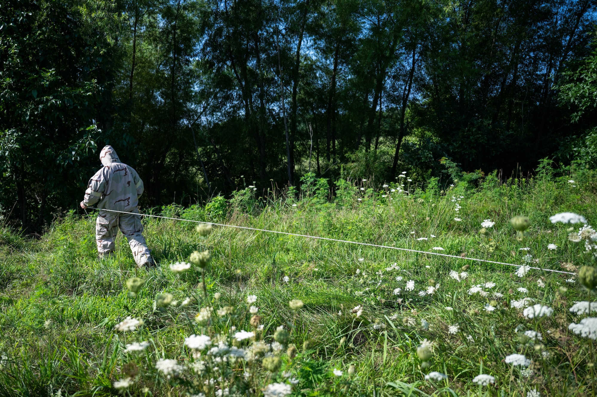 A man in MOPP gear walks with a line towards the trees