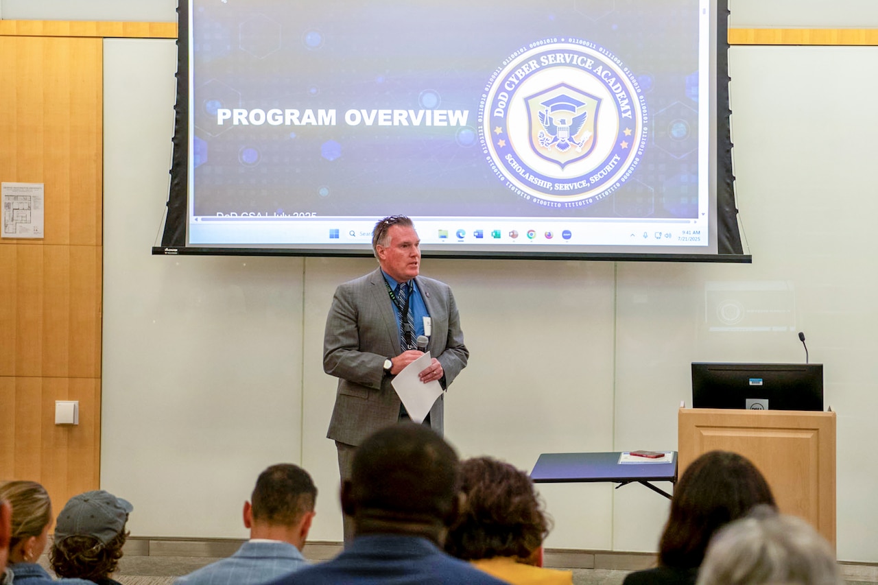 A man wearing a business suit stands in front of a room with a microphone. There is a projection screen behind him and a group of people in front of him.