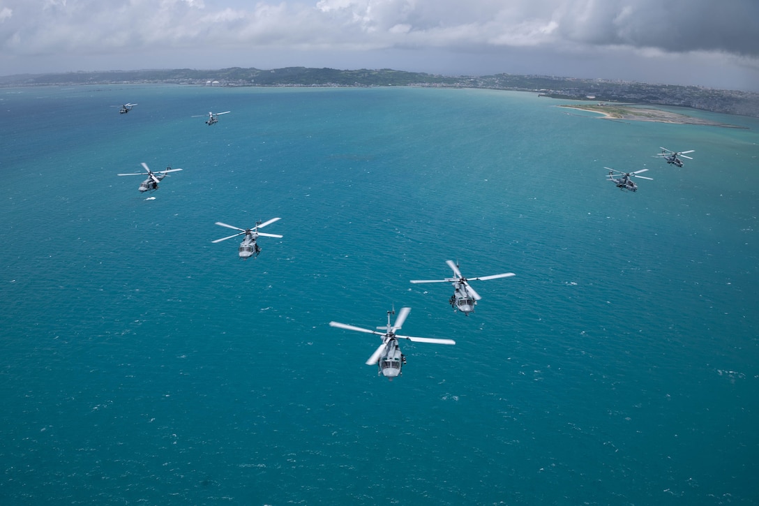 Eight helicopters fly over the ocean during the day, as shown from above.