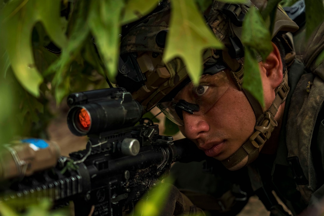 A close-up of a soldier in tactical gear aiming a service weapon at night under cover of leaves is shown.