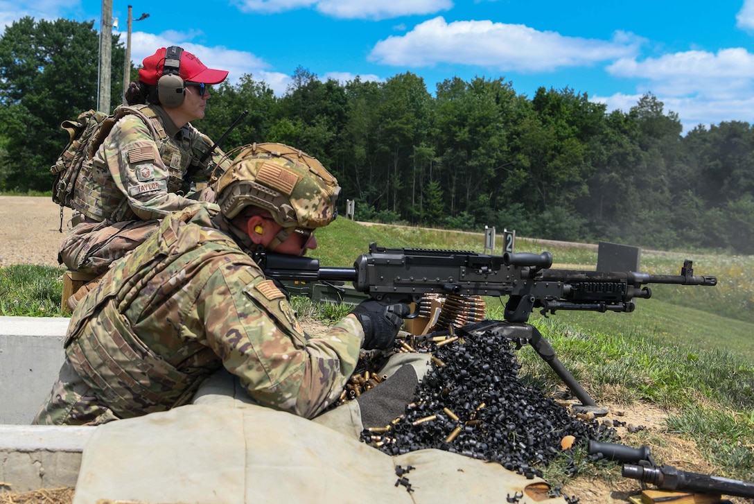 A soldier in tactical gear fires a machine gun from the grass near a forest while another soldier watches.