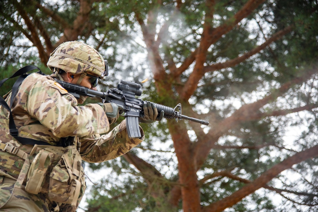 A soldier in tactical gear fires a service weapon outside with a tree in the background.