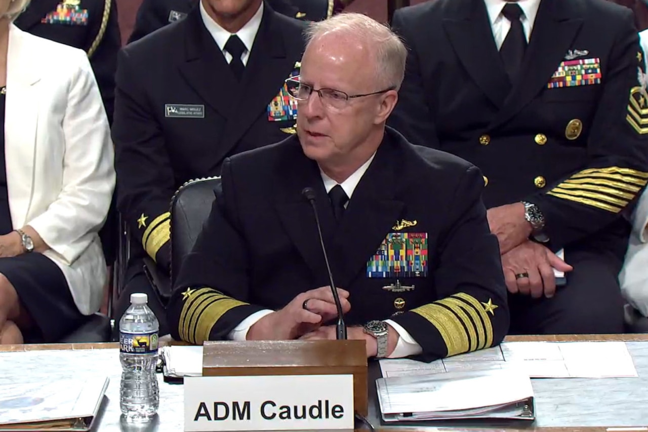 A man wearing a formal military uniform sits behind a desk and speaks into a microphone, with other people seated behind him.