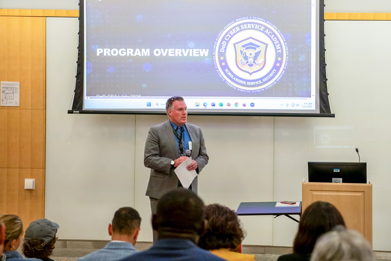 A man wearing a business suit stands in front of a room with a microphone. There is a projection screen behind him and a group of people in front of him.