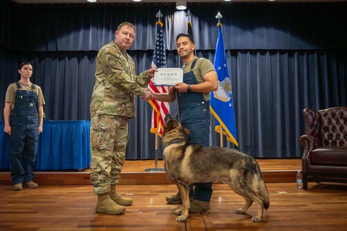 Two U.S. Air Force Airmen hold a certificate with a military working dog stands on stage.