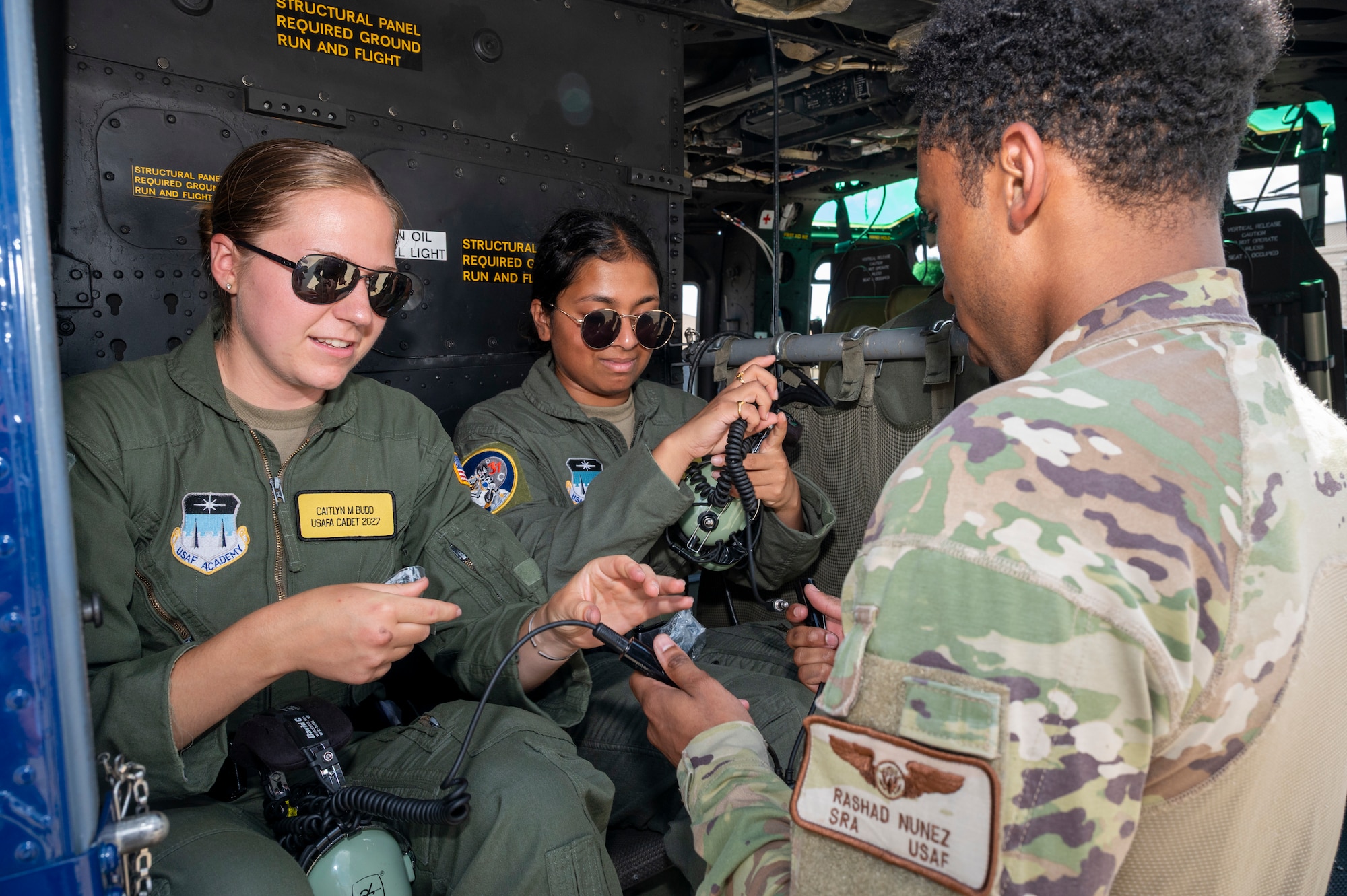 U.S. Air Force Senior Airman Rashad Nunez, right, a special missions aviator with the 1st Helicopter Squadron, assists U.S. Air Force Academy Cadets Caitlyn Budd, left, and Keerthanaa Narasingu don headsets at Joint Base Andrews, Md., July 17, 2025. Cadets visited JBA and Joint Base Anacostia-Bolling, Washington, D.C., as part of USAFA’s Summer Operations program to learn more about officer life and the operational Air Force. (U.S. Air Force photo by Hayden Hallman)