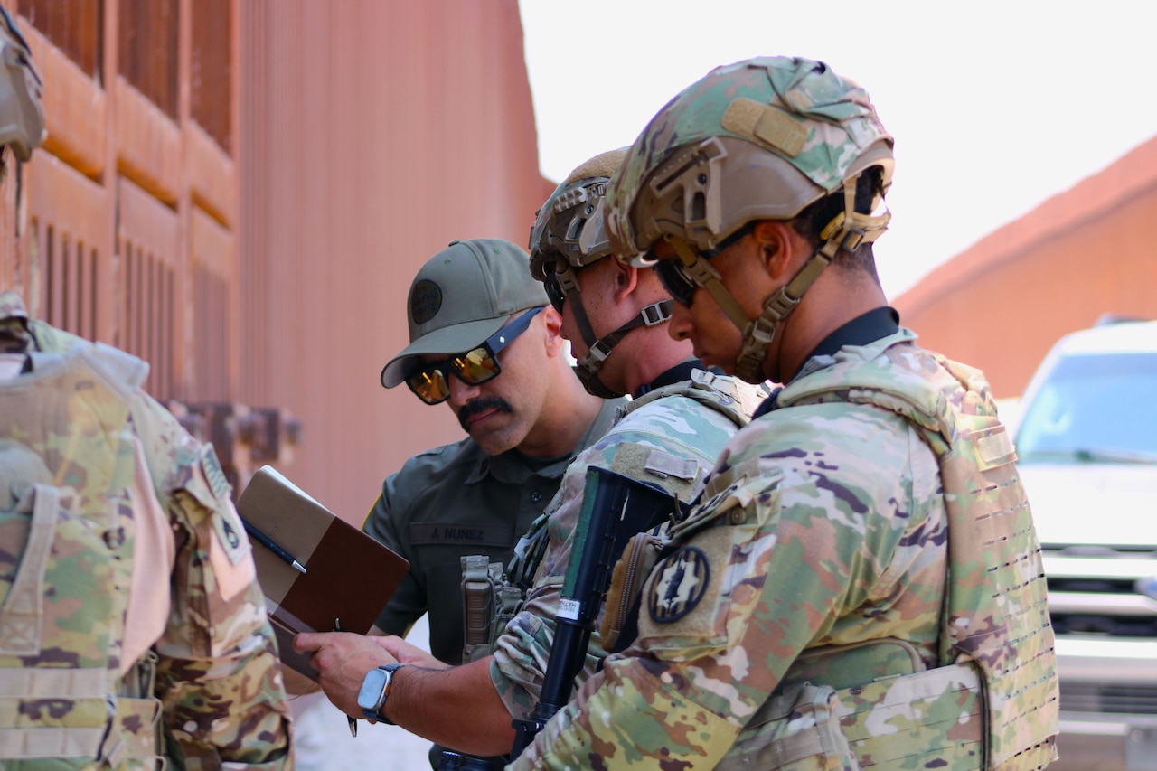 Two soldiers speak to a person in a baseball cap outside, with a large structure and vehicle in the background and another person nearby.