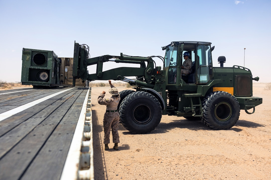 A Marine gestures next to a tractor outside in a desert setting under a cloudless blue sky during the day.