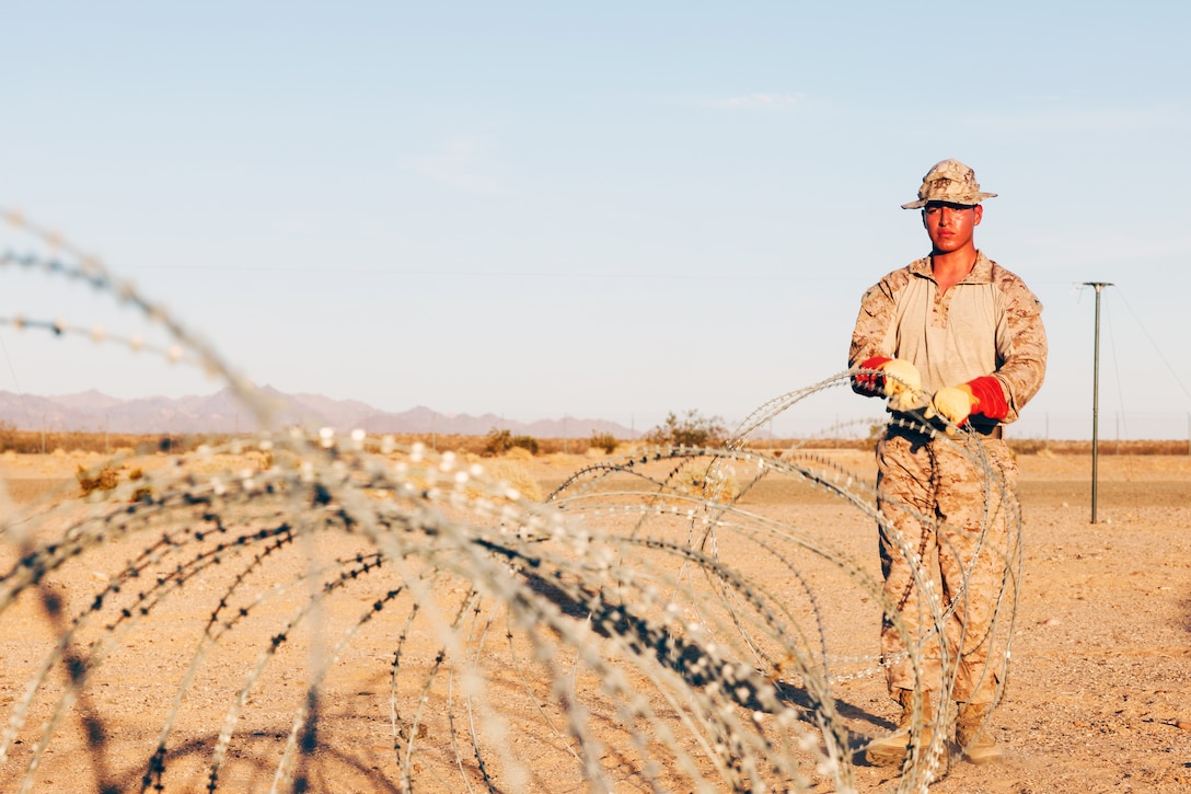 A Marine stands in a desert setting and places concertina wire on a cloudless, sunny day.