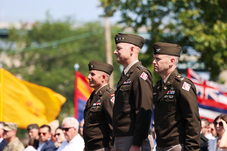 Three men in Army officer uniforms stand at attention outdoors while an audience observes.