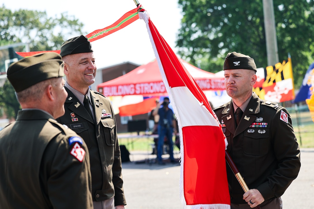 Three men stand outdoors in Army officer uniforms. One of the men holds a military flag.