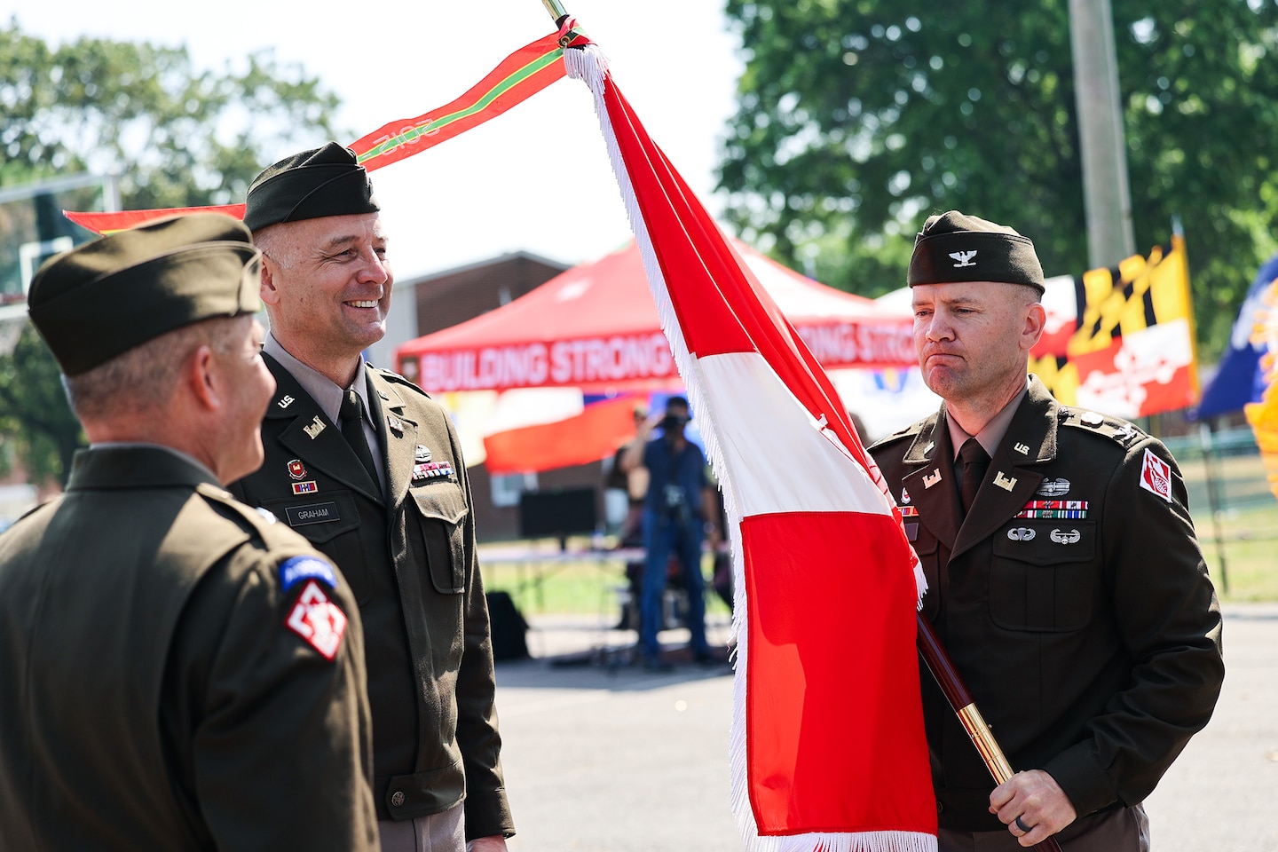 Three men stand outdoors in Army officer uniforms. One of the men holds a military flag.