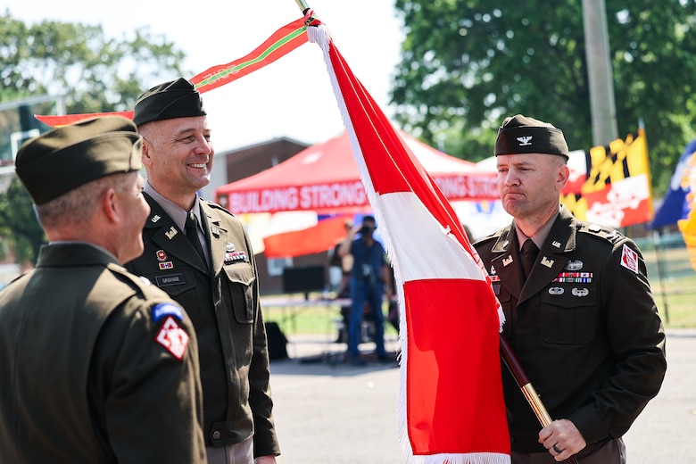 Three men stand outdoors in Army officer uniforms. One of the men holds a military flag.