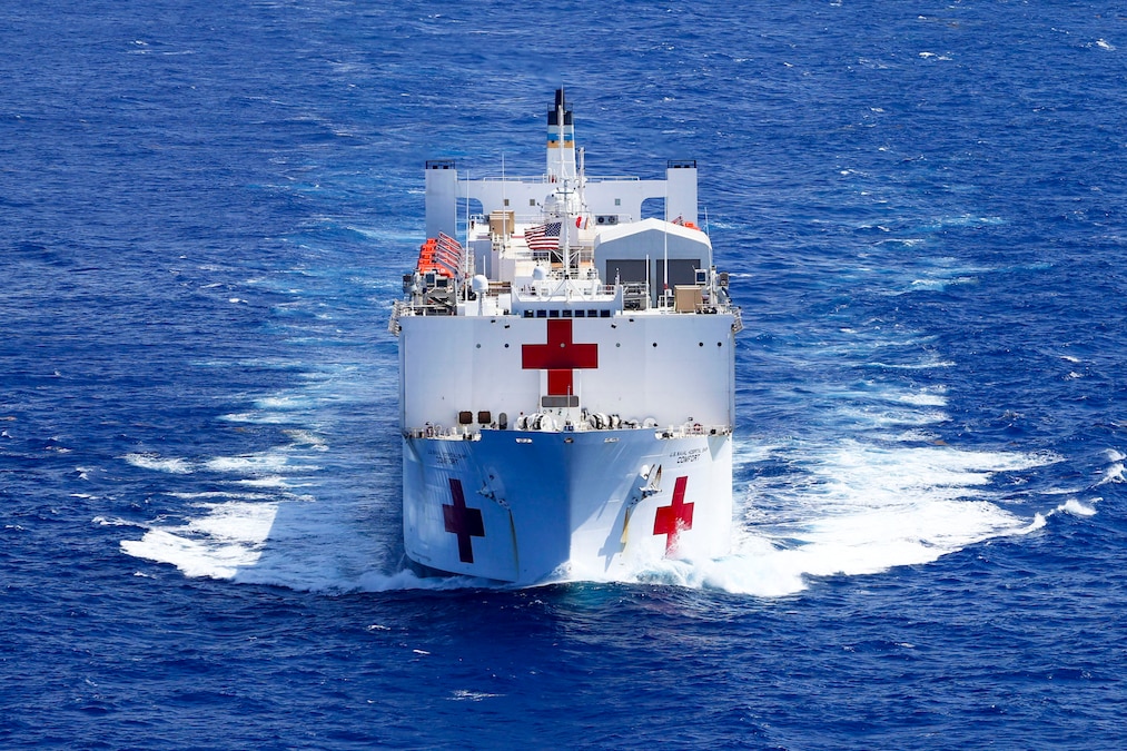 A medium close shot of a white ship with three red crosses and sailors working on deck as it travels through blue waters.
