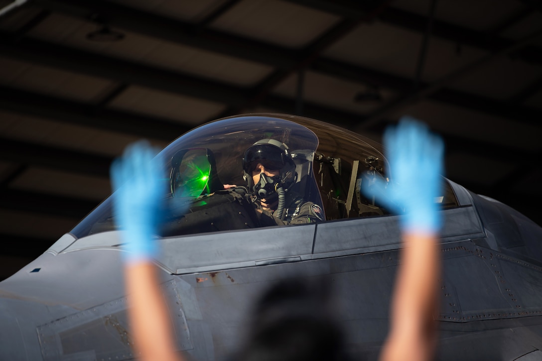 An airman sits in a cockpit against a dark background as a fellow service member wearing blue gloves raises their hands in the foreground.