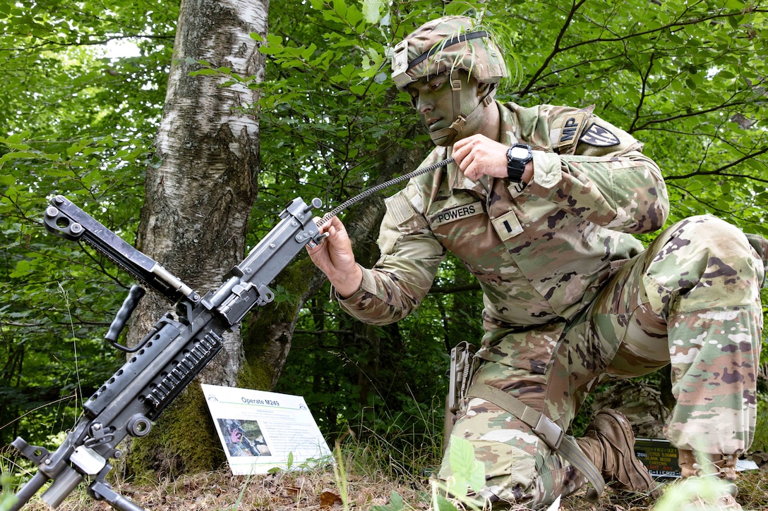 A soldier wearing camouflage kneels in the woods while disassembling a weapon as a sign rests on a tree.