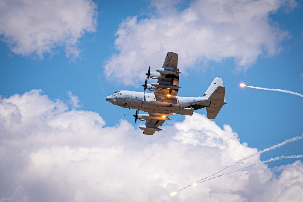 An aircraft flies against a cloudy, blue sky as flares descend from behind as seen from below.