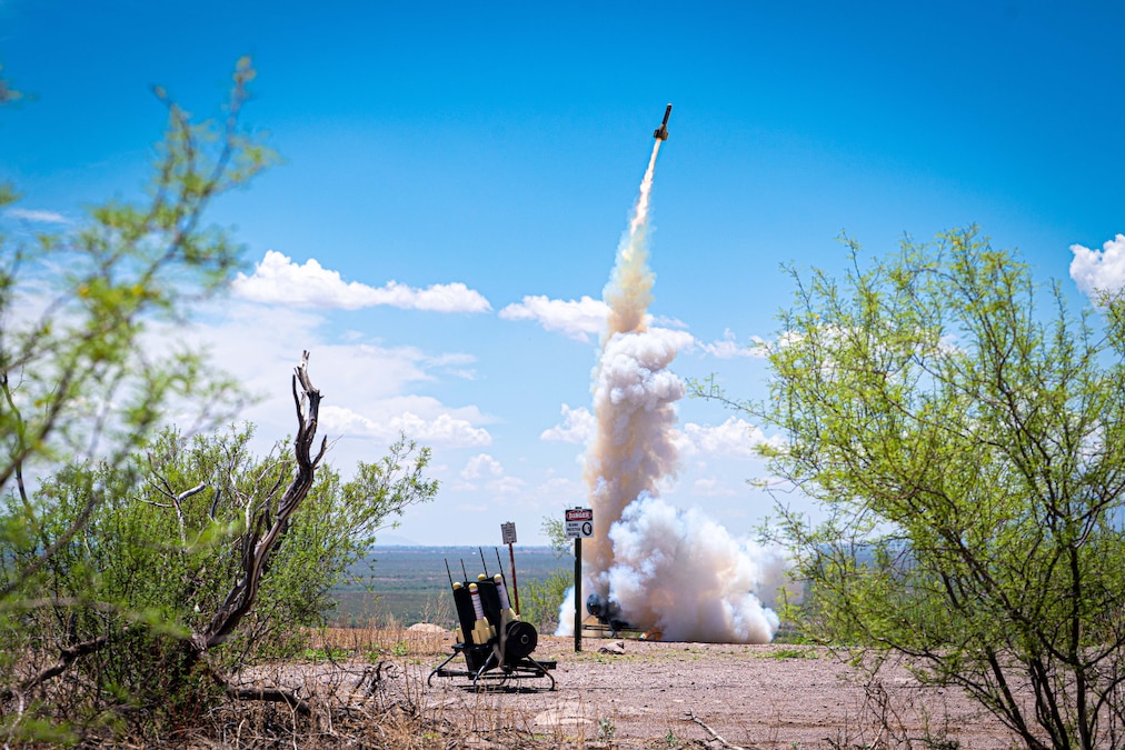A rocket leaves behind a trail of smoke after lifting off into a blue sky from a field in between bushes.