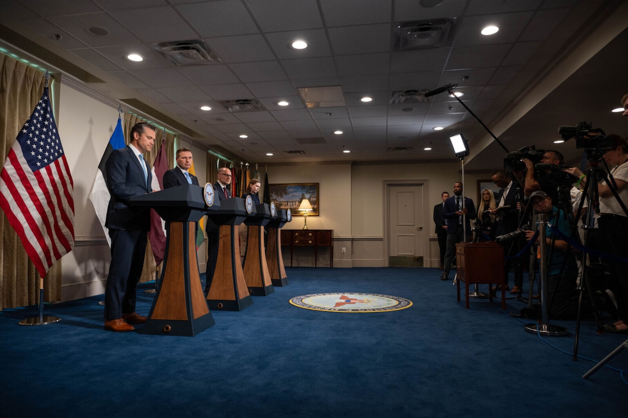 Four individuals wearing business attire stand behind lecterns. Behind them are flags representing multiple nations. Before them are journalists with video cameras and still cameras.