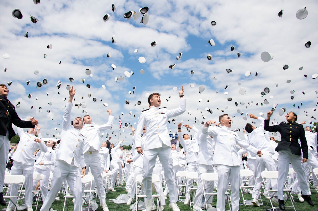 Service members in white uniforms and black uniforms toss their hats into the air.