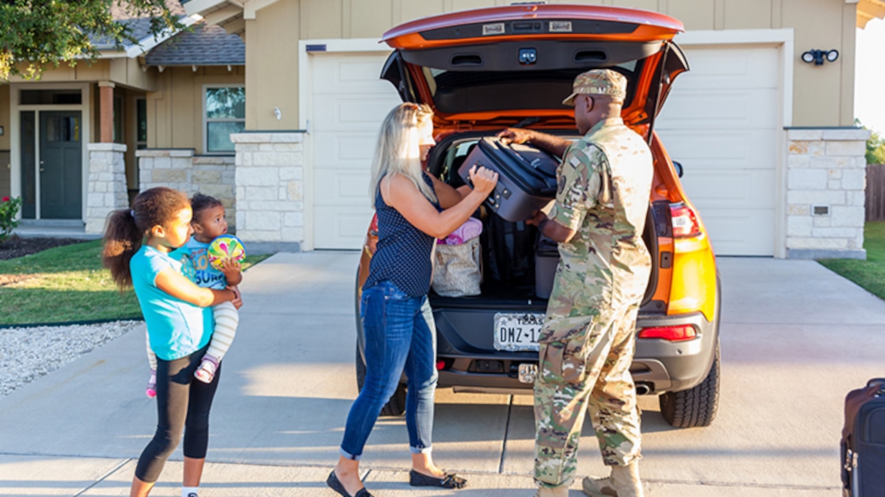 A man and woman unload a suitcase from a car as a child holding a baby watches.