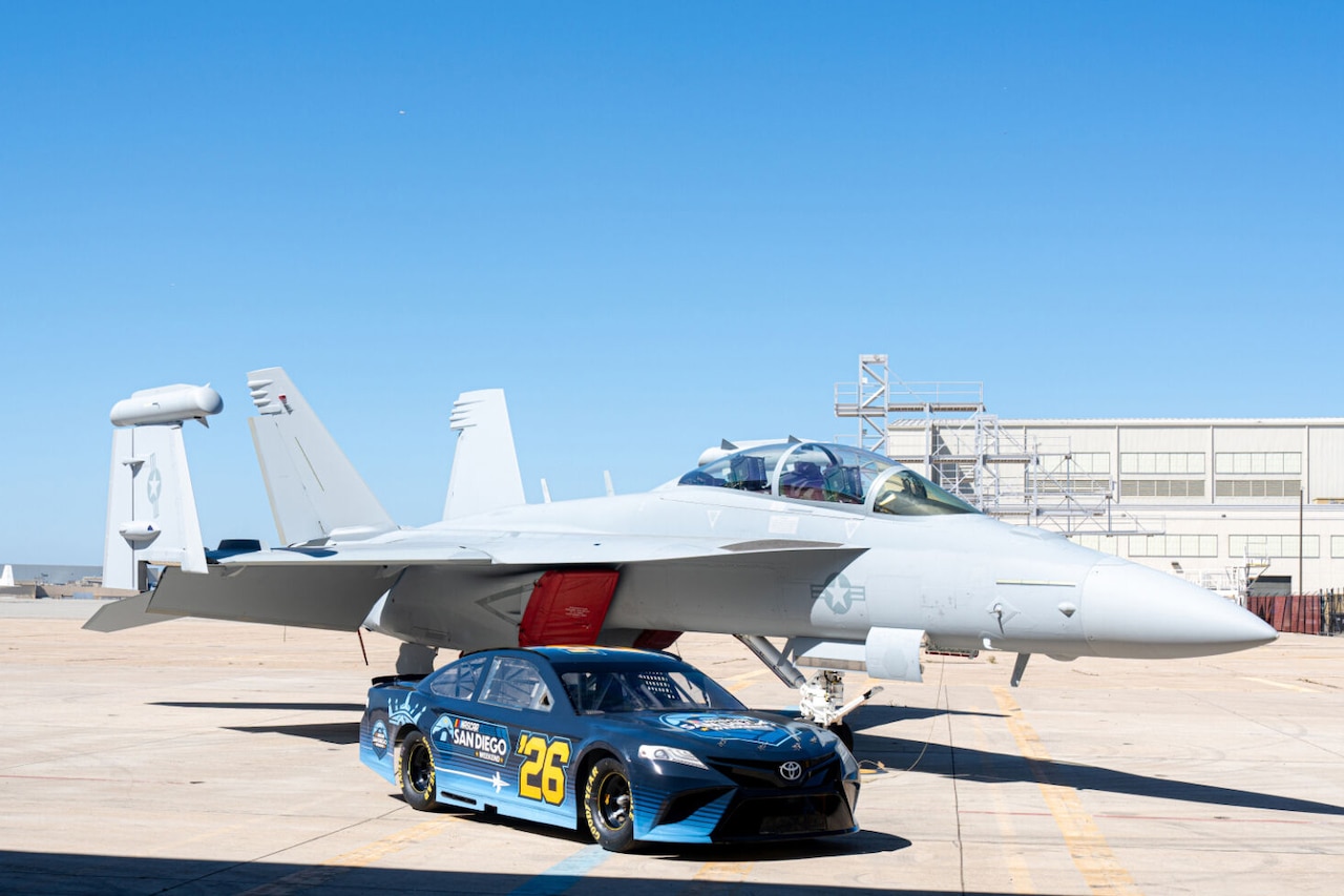 A race car sits in front of a fighter aircraft.