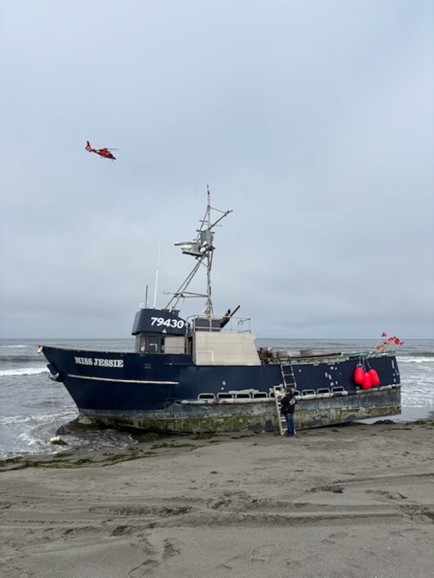 A Coast Guard MH-65 Dolphin helicopter is seen flying behind a beached fishing vessel.