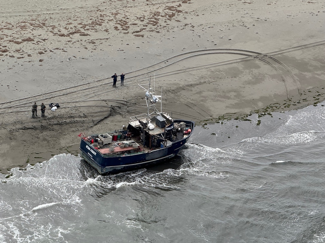 An aerial shot of a beached fishing vessel with one person on the vessel and another 5 standing on the beach behind it.