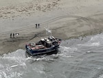 An aerial shot of a beached fishing vessel with one person on the vessel and another 5 standing on the beach behind it.