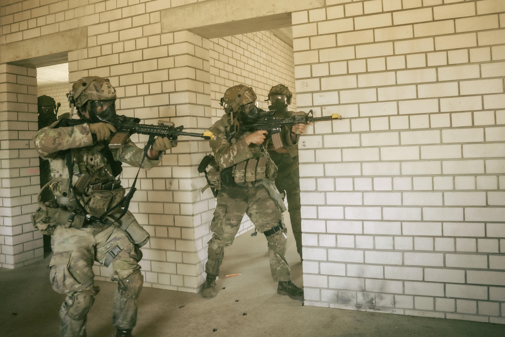 Soldiers in combat uniforms point rifles beside a white brick structure.