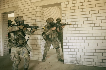 Soldiers in combat uniforms point rifles beside a white brick structure.