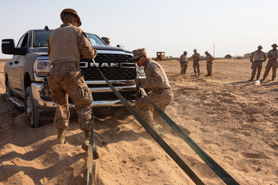 Two Marines attach straps to the front of a truck stuck in desert-like sand on a sunny day as others stand in the background and watch.