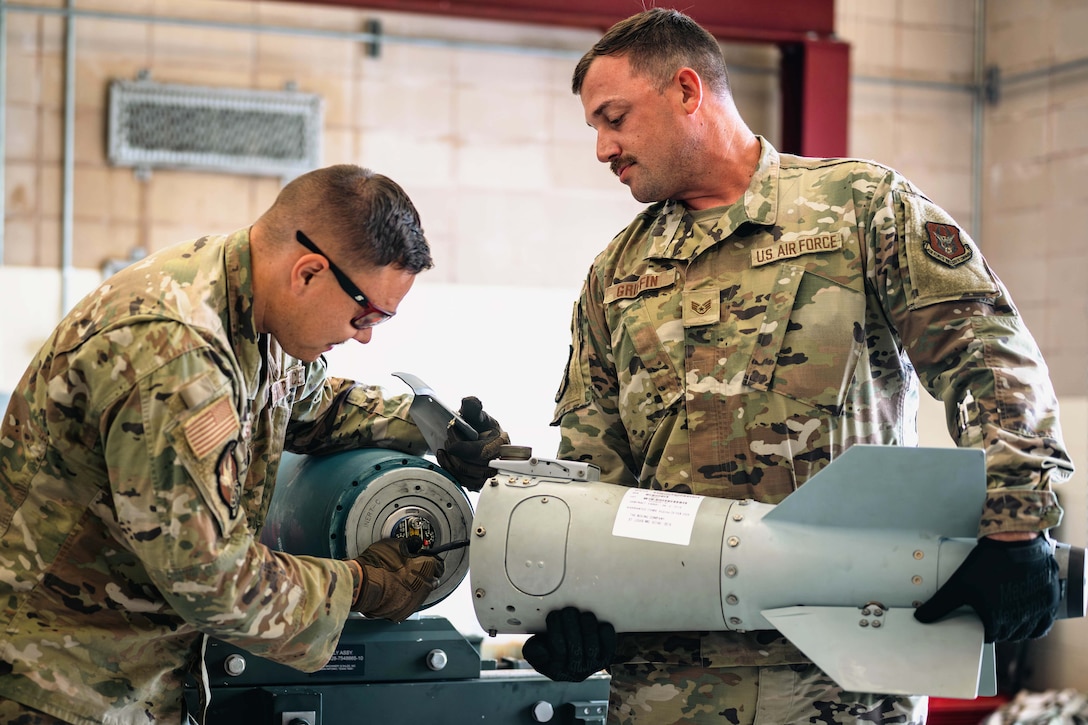 Two airmen wearing protective gear connect two pieces of a weapon in a garage-like area.