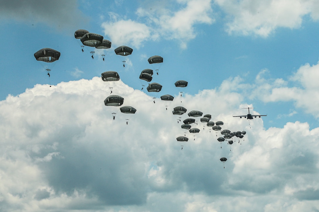 An aircraft flies in the distance in a cloudy, blue sky as dozens of soldiers descend with parachutes behind it.