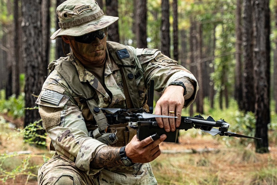 A soldier in camouflage kneels in the woods while holding a drone.