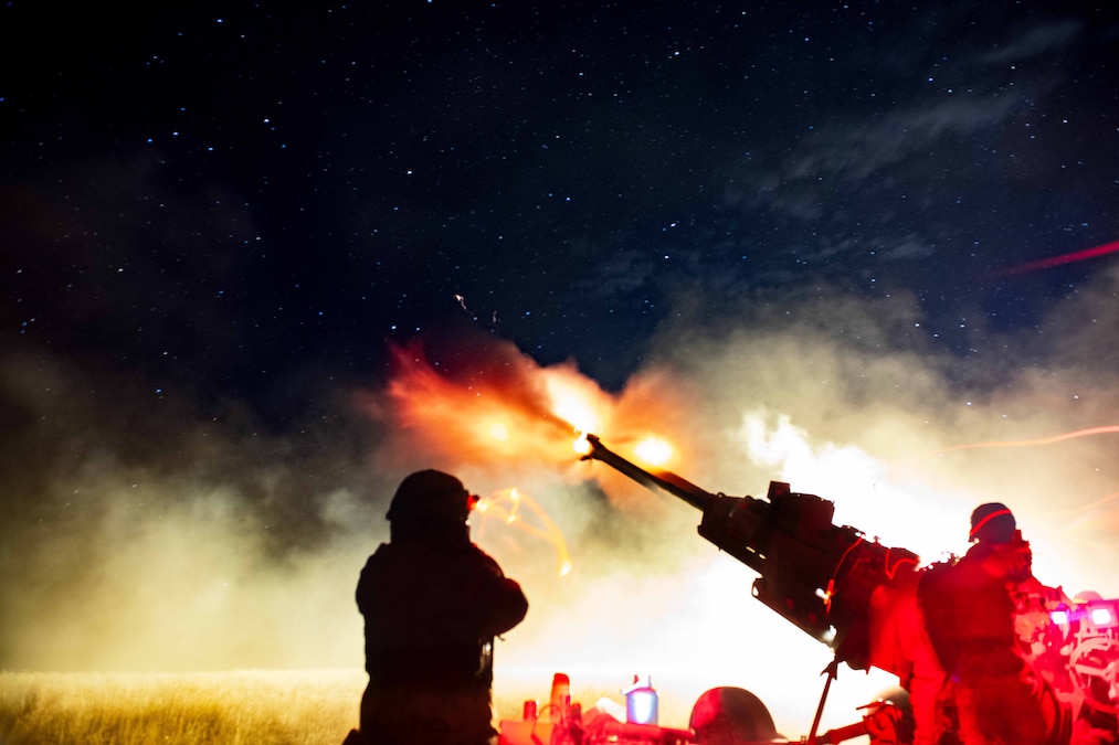 Soldiers in silhouette surrounded by smoke fire a weapon creating an orangish blast against a starry sky illuminated by red streaks.