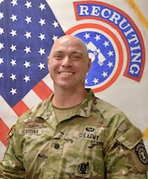 Male Soldier smiles while standing in front of the American flag and the US Army Recruiting Command flag