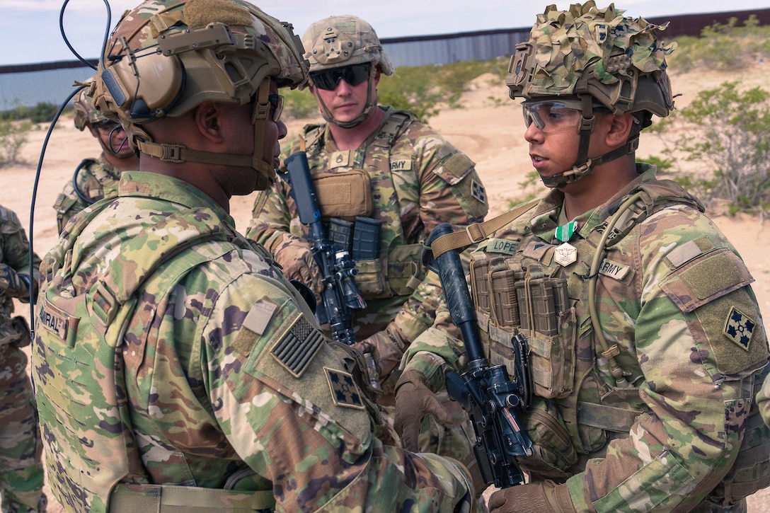 A soldier in combat gear prepares to shake hands with another soldier as others looks on while standing in a sparsely vegetated area with a wall in the background.