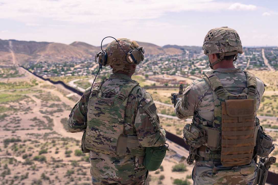 Two soldiers, one gesturing with a hand out, look from an elevated position over sparsely vegetated land divided by a wall, with hills in the distance.