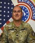 Soldier smiles while posing in front of American flag and United States Army Recruiting Command flag.