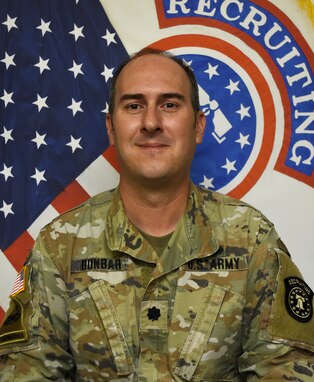 Soldier smiles while posing in front of American flag and United States Army Recruiting Command flag.