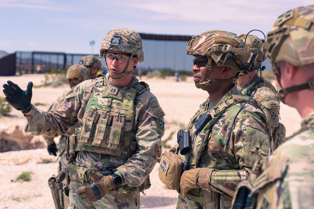 A soldier gestures with one hand while speaking to another soldier who is looking into the distance as others stand near them on sparsely vegetated land, with a tall, fence-like structure in the background.