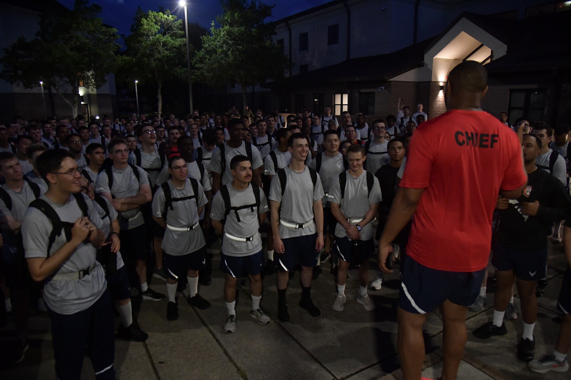 Airmen standing in a circle listening to a chief in a red shirt speak.