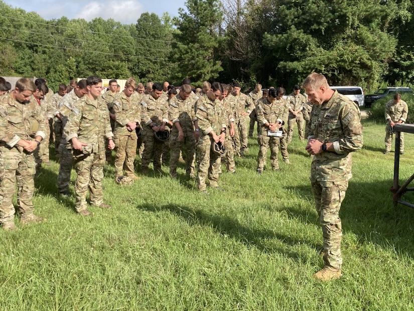 More than a dozen uniformed soldiers stand outside and pray. A man dressed in a military uniform, stands in front of them. All of their heads are bowed.