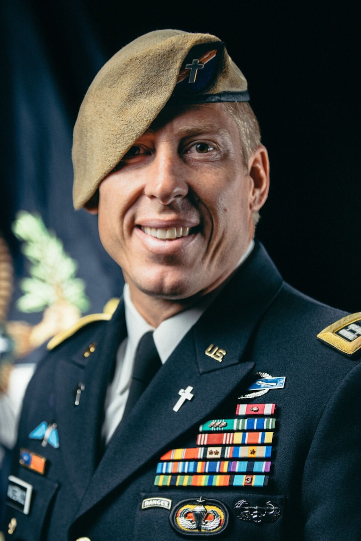 A man dressed in a military uniform wears a beret adorned with a small cross.