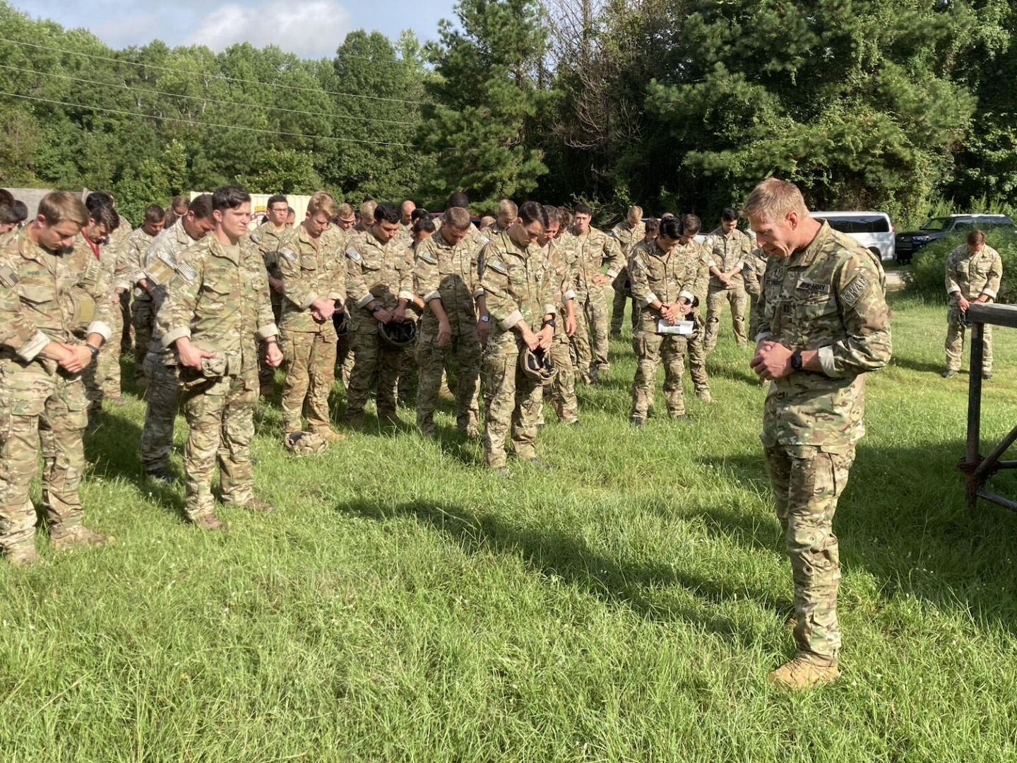 More than a dozen uniformed soldiers stand outside and pray. A man dressed in a military uniform, stands in front of them. All of their heads are bowed.