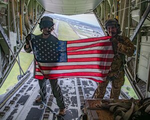 U.S. servicemembers display an American flag over an airfield out of the rear of an aircraft.